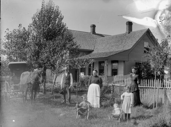 Group portrait of people posing near a picket fence in front of a frame house. On the left is a single horse with a fly-net pulling a buggy. A man with a beard and holding a hat is standing nearby. A woman wearing an apron is standing in the center. In front of her is a young child sitting in a low chair, and a young girl with a doll in small baby carriage. Behind her a young woman wearing a dark dress is standing in front of the picket fence.