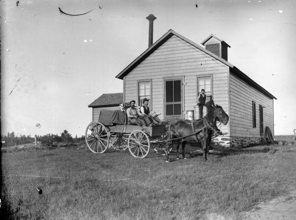 Two men are posing sitting in a wagon pulled by a team of two horses. One of the men is holding the reins, and the other man is holding tools. Behind them a man is sitting among metal containers on an open porch or loading dock of a building, perhaps a creamery, which has a tall metal chimney.