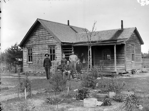 Seven people are posing in the yard of a frame house with a porch. There are plants in a garden in the foreground. An older man and woman are sitting in chairs, and behind them are standing are two men, two young girls, and a young boy. The men and young boy are wearing hats.