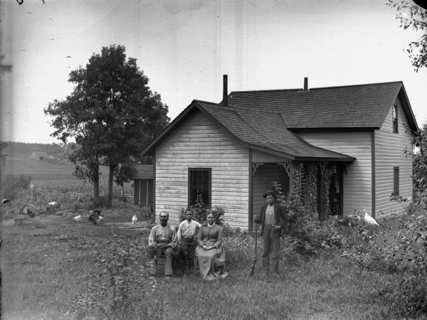 Group portrait of a man and woman posing sitting in chairs, with a young boy and a dog posing between them. An older boy is standing to their right holding a gun. In the yard are chickens and turkeys, and behind them is a frame house with a porch. In the far background on the left are fields, and what appears to be another farm with farm buildings.