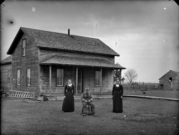 View across yard towards a man sitting in a chair, with two woman standing on the left and right. The two women are wearing black dresses with white bows tied at their necks. The younger woman on the left has hair clips in her hair. The man is wearing work clothes, and his hat is beneath his chair. Behind them is an unpainted frame house with porch and wooden walkway. Farm buildings with fence and fields are in the background on the right.
