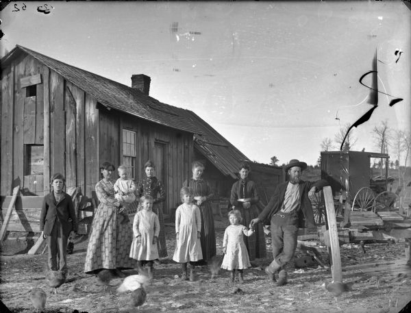 Group portrait of a man, four women, an infant, three young girls, and a boy in a yard with chickens and cats. In the background on the right is a photographer's wagon.