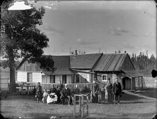 Two groups of people are posing in yard. On the left a man and woman are posing sitting, with the man resting his hand on a dog. Standing around them are three young girls, and a young boy. On the right are two women standing along with a man, and a young girl. The woman in the middle is holding a small dog. The dwelling in the background is a combination of log construction with a rough board addition.