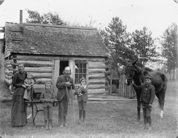Standing in the yard in front of a log house is a man standing and holding crutches in the middle, a woman on the left holding a small girl who is perched on a sewing machine table, and two young boys, one of whom is holding a cat. On the right an older boy is standing holding the reins of a horse. Behind them standing in the cabin and looking out the window of the house is a young girl, with possibly another child next to her.