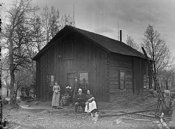 Group of adults and one infant in front of a house with chink-log construction along one side, and board siding on the front. On the left a woman is standing next to another woman who is wrapped in a blanket and sitting in a rocking chair in front of a door. Next to her is standing a man wearing a hat. In front of him an older man and woman are sitting in chairs. The woman is holding an infant in her lap. In the foreground is a rooster, and an axe resting on a log.