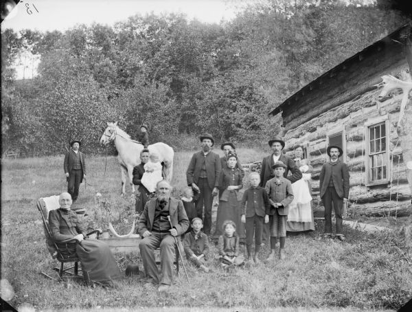 Large group of people posing with a horse in the yard next to a house with chink-log construction. From the left in front is an elderly woman sitting in a rocking chair next to an elderly man, who is also sitting. In the center is a young boy and girl sitting on the ground, and next to them on the right are two older boys standing. Standing left to right behind the first group is a woman holding an infant in her arms, a man standing wearing a hat and holding the hand of a young child, a man and woman, and then two men standing just in front of a woman who is holding an infant in her arms. In the background on the left is a man is standing holding the reins of a horse on which a young boy is sitting bareback.