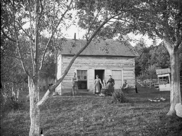 View across lawn of group posing in front of a log house, which has two large windows on either side of the open door. A man and woman are sitting in chairs, and standing with them are two young women and a young man. There is a well on the far right. Probably a farm on County Road X, quarter of a mile east of the Trout Run Methodist Church, on the way to Melrose.	Note on back of print reads, in part: "1/4 mile of east of Trout Run Methodist Church, on the way to Melrose. The place used to belong to a party named Stratton."