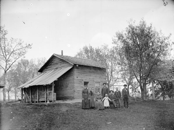 View across yard towards a group of eight people posing in front of a log house. On the left two young women are standing near an older woman, who is sitting in a chair. A young girl is wearing a white dress and is standing in the center, and on her right is an older man sitting and smoking a pipe. On the far right two young boys are wearing hats are standing. Behind the group an older man is standing wearing a hat. Under an open sided roofed area on the left side of the house, corn is hanging to dry.