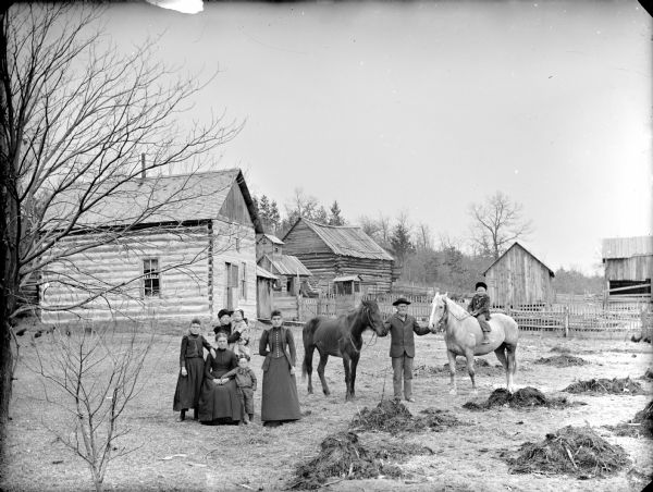 Group posing in yard with log buildings in the background. In the center a woman is sitting, and a young girl and small boy are standing next to her. In the background, a woman is standing and holding an infant in her arms. A woman is standing nearby, and on the right an older man is standing holding the reins of two horses, with a boy mounted bareback of the lighter colored horse.