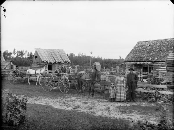 Group portrait of a man, woman, and four children posing standing in front of a wooden fence. On the far left is a horse pulling a small buggy, a young girl and two young boys are in another horse and buggy, and on the right a man and woman are standing with a young girl. In the background is a log house, farm buildings, and a birdhouse on a tall post.