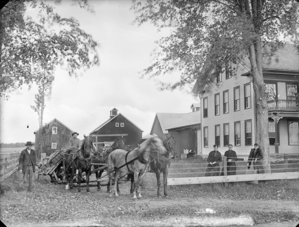 Man posing sitting on farm equipment, probably a grain-binder, pulled by a team of four horses in the driveway of a large two-story frame house. A man is standing on the left near a fence. Three women are posing in the yard on the right, standing behind a wire and wood fence. Farm buildings are in the background.
