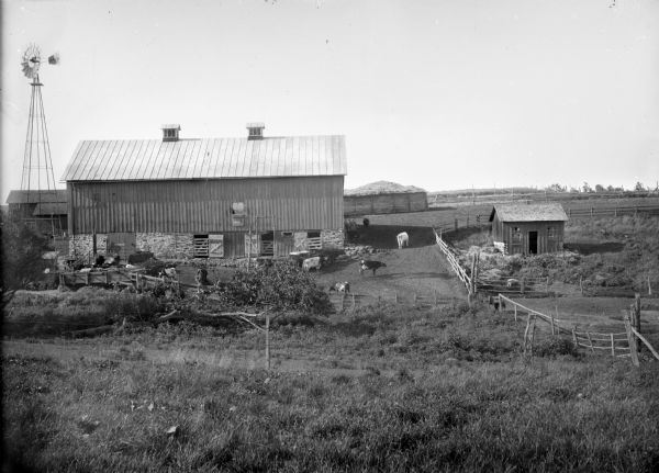 Elevated view from hill of farm buildings, barn, windmill, and cows in a pasture surrounded by a wooden fence. A sign on the barn door reads: "No Smoking."