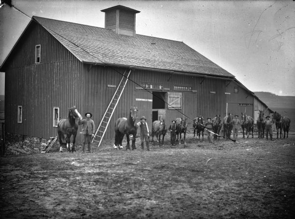 Group portrait of six men displaying ten horses in front of a barn, possibly the Bright-Withel farms near Hixton.	
