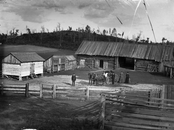 Elevated view over fences towards a yard surrounded by log farm buildings. In the yard posing are two women, two girls, and a boy sitting in a wagon pulled by a team of two horses. A man wearing a suit and hat is standing next to the wagon holding the reins of the horses. Near him on the right are standing two women and a young girl. Another man is standing near the horses on the left. There is a field, and a hill with trees and shrubs, in the background.