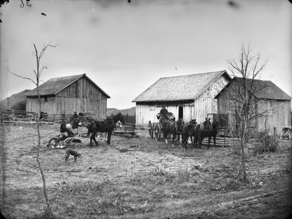 In the center is a man posing sitting on a plow pulled by a team of two horses. In front of him is a dog, and another dog is lying behind the plow. Standing behind is a woman standing holding a basket or pail near a flock of chickens in front of a parked wagon. On the right a man is sitting in a fur-covered wagon seat holding the reins of a team of four horses. A covered carriage is parked near a small building on the right, and two more barns and a haystack are behind the group. In the far background are trees and hills.