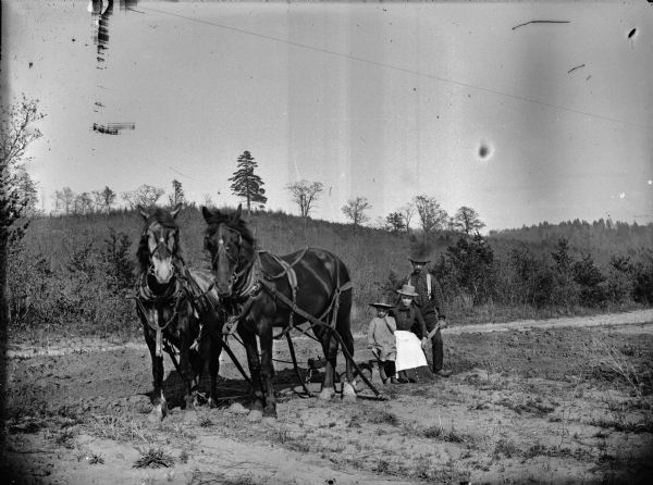 A woman and child are posing sitting on a plow steered by a man standing on their right. The two horses are standing on the left. In the background is a tree-covered hill.