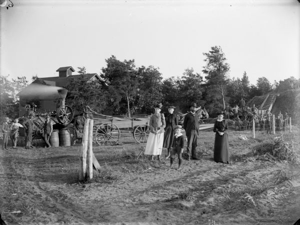 People posing in field, with a barn in the background among trees. In the center a man, three women and a boy are standing in front of a wire fence. Behind the group is a horse and wagon. On the far left several men and boys are operating a steam tractor that is being used for belt-driven threshing machinery, which is surrounded by a large group of men and boys near a large haystack on the far right.	