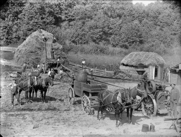 Elevated view of men and boys operating a steam tractor used for belt-driven threshing machinery. Farm buildings are on the right, and one has a thatched roof.	
