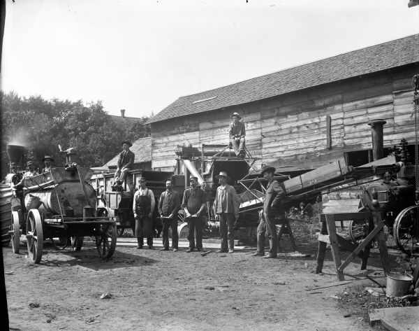 Large group of men posing standing by and sitting on farm machinery, including a thresher and steam tractors. Probably at the Thompson Foundry and Machine Shop, located along Town Creek for water power.