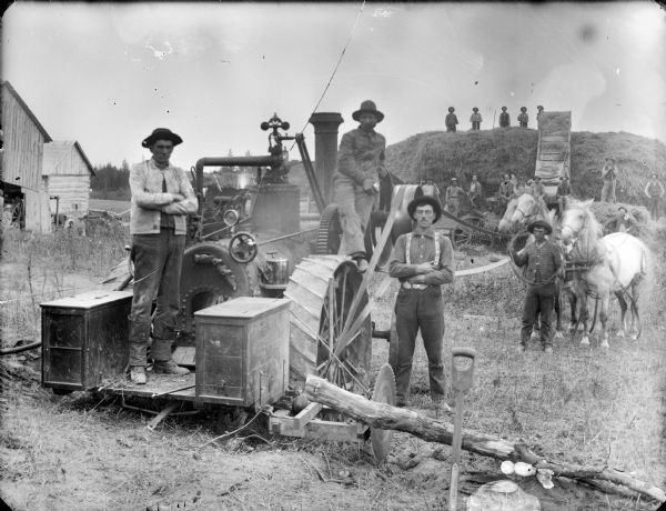 A group of men are operating a steam tractor and threshing machinery near farm buildings. Three men are posing in the foreground near the tractor, and another man is standing behind them with two horses. A large group of men are posing in front and on top of a large stack of hay in the background.