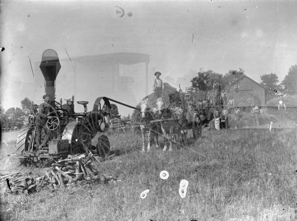 Men operating a steam tractor and threshing machinery near farm buildings. The negative is double exposed and shows the outline of a photographer's wagon.	