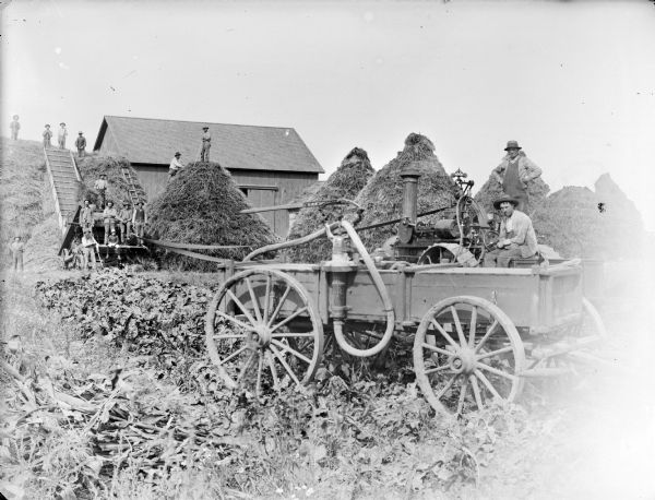 Men operating a steam tractor and threshing machinery near farm buildings. Two men are posing on the right with the tractor. On the left a group of men are posing near a tall stack of hay.