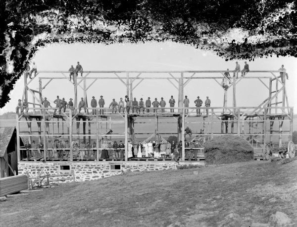 Group portrait of a large group of workers, along with women, children, and dogs, posing on the frame of the barn they are building.