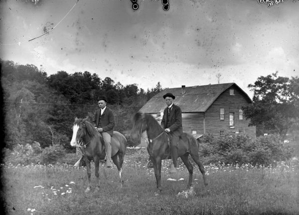 Two men are posing mounted on two horses in a field in front of a wooden building, probably the Charter Oak Mill.	