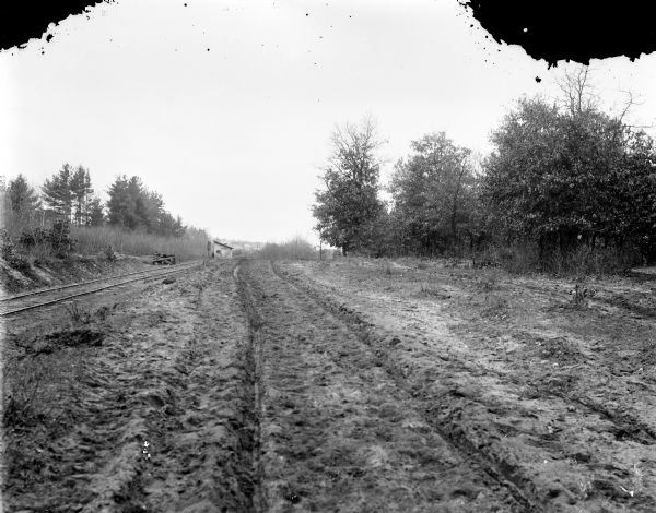 Rutted, dirt road running along railroad tracks toward a small wooden building. In the far background is a cluster of houses.
