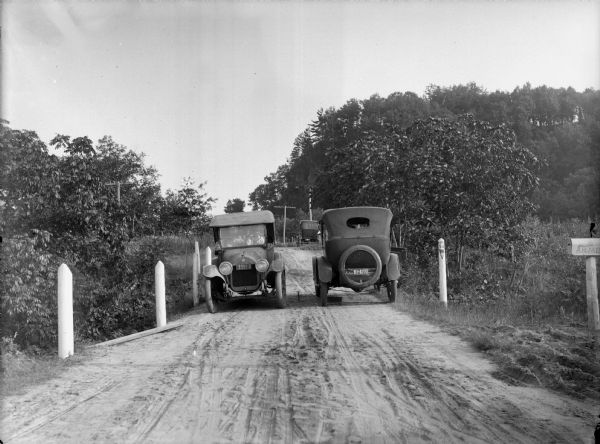 View down dirt road towards two automobiles, next to each other and going in different directions. Another automobile is parked in the background. On the right is a mailbox, and near it is a white wooden post at the side of the road marked: "52 Wis." In the background is a tree-covered bluff.
