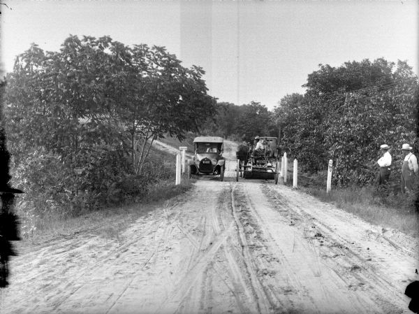 View down dirt road towards a man, seen from behind, sitting on a plow pulled by a team of two horses, near an automobile facing the camera. Next to the automobile is Al Meek's mailbox. Another automobile is parked at the side of the road in the background on the right. Two men are standing at the side of the road on the right next a wooden post with a painted sign: "52 Wis."