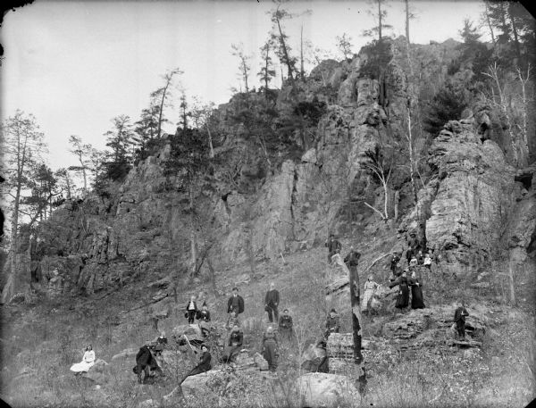 Group of men, women, and children posing sitting and standing among the rocky outcrops at the base of a rocky hill.	