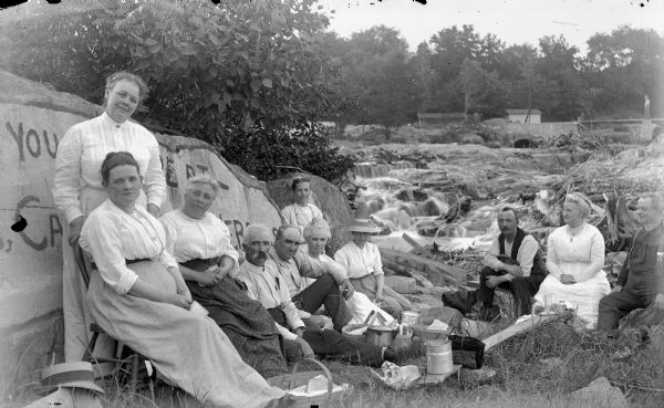 Four men and seven women are posing sitting and standing around a picnic on the edge of a river, with a man standing on a bridge in the background. The first man on the left is probably Eugene Greenlee. A large rock on the left beside the group has a painted sign.