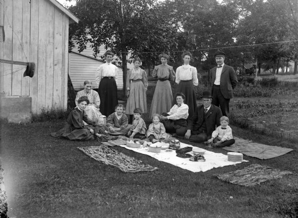 Five children, three women, and a man are posing sitting, and four women and a man are standing, around a picnic in a yard in front of wooden buildings. Probably the R.P. Rainey and S.E. Vance Families.	