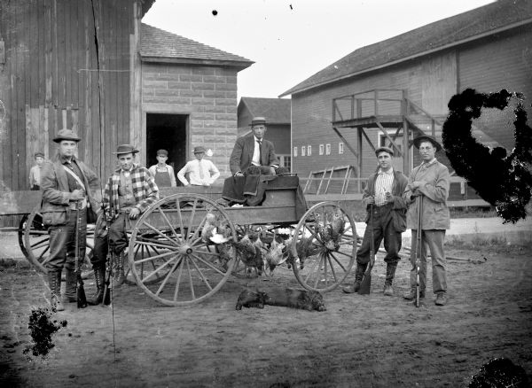 Group of people posing with a wagon from which several dead prairie-chickens are hanging. One man is sitting in the wagon with a dog. Seven men are standing around the wagon, with four of them holding shotguns. A dog is lying on the ground in front of the wagon. In the background are wooden and brick buildings. Identified men in the foreground are, left to right, Fred Werner, Will McGillivray, Dr. A.S. Trondson (in the wagon), unidentified man, and Albert McDonald.	