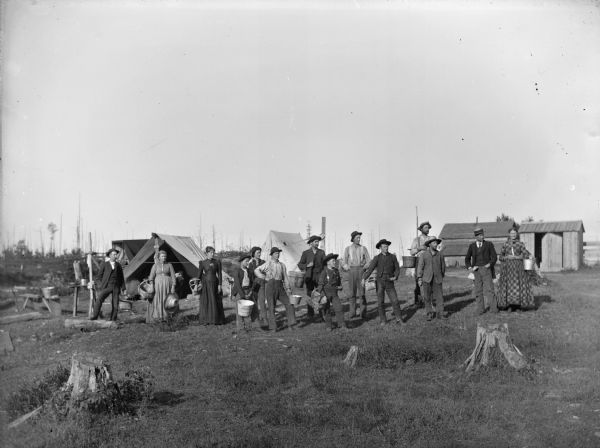 Eleven men and four women are posing standing in front of tents and farm buildings. They are holding buckets and baskets. 	
