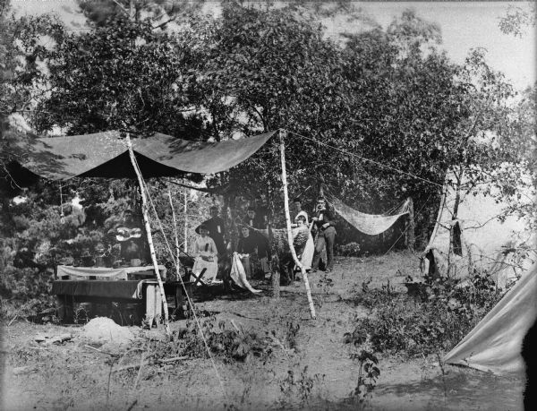 Men and women are posing standing and sitting under a rough awning. They are sitting in chairs and a hammock near a table in the woods. A few people are wearing ribbons pinned to their chests.