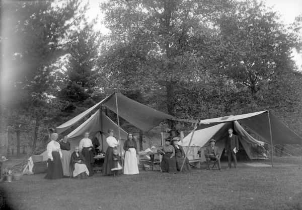 Six women, two men, and a girl are posing sitting and standing near a table under the awning of two tents in a clearing in the woods.	