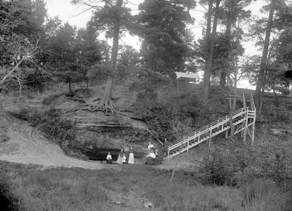 Seven women, a dog, a young girl and a man are posing standing at the bottom of a rocky hill. A steep stairway is leading up to the top of the hill, where a tent is set up. Probably at Rock Spring Park.