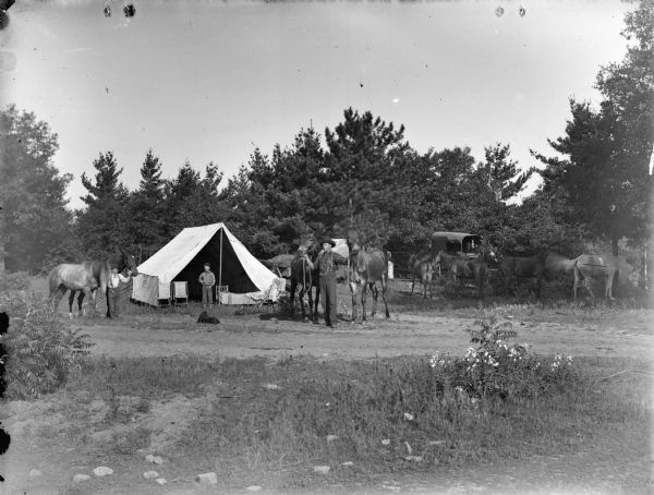 View across dirt road towards two men standing, each holding two horses, on either side of a tent. A boy is standing in the center in front of the tent, and in front of him a dog is lying on the ground. On the right four horses are grazing in the grass near a photographer's wagon.	