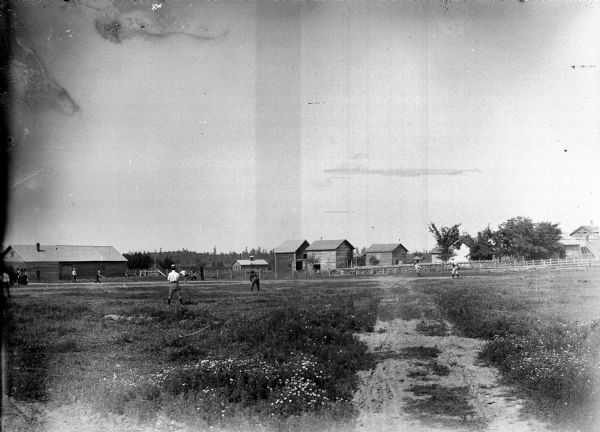 Men are playing baseball in an open field in front of farm buildings and a small group of people.	