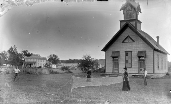 Two men and two women playing lawn tennis. Behind them is the front of a small church, a large two-story house and other wooden buildings in the far background. The church possibly became the Occa Feeds Warehouse, No. 74, on Highway 54.