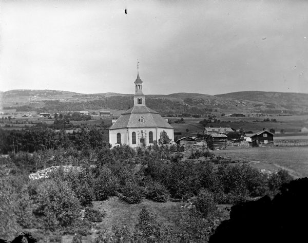 Copy photograph of an elevated view of a hexagonal building, possibly a church, in a countryside with several other buildings in the landscape. Probably the Sor-Fran Church in Gudbrainldalen, Norway, looking across Langue toward Oppland; many individuals in the Black River Fall area came from this region.