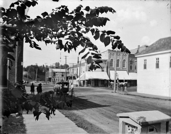 Elevated view over sidewalk of the intersection of Main and First Streets. The limbs of a tree are in the foreground. On the opposite corner is the Palace Bakery. Pedestrians are standing near a horse-drawn buggy. In the foreground near the sidewalk is a display case with photographs.