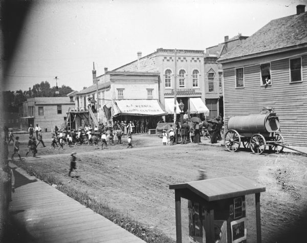 Elevated view over sidewalk of the intersection of Main and First Streets, towards storefronts including A.F. Werner, Leading Clothier. A crowd is gathered, probably watching a parade. In the foreground is a roofed display case of Van Schaick's photography.