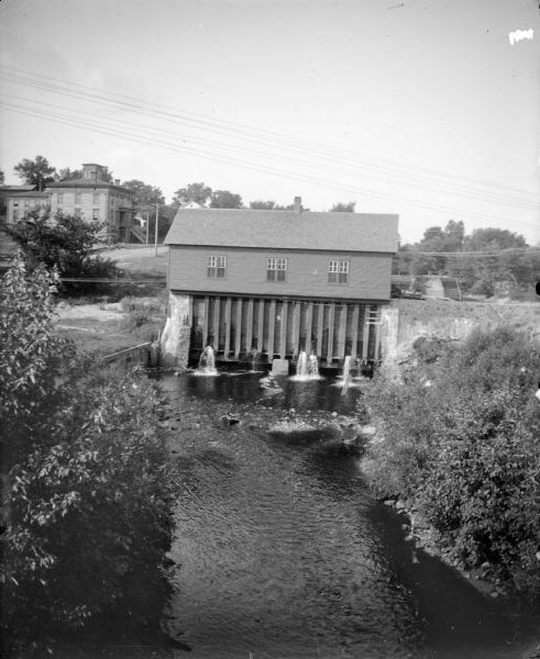 Elevated view of the river dam and what is probably the powerhouse. Trees and bushes line the riverbank in the foreground. In the background on the left is a road, and a large three-story brick building.
