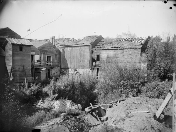 Wooden buildings in ruin. Overgrown bushes and piles of dirt are in the foreground.	