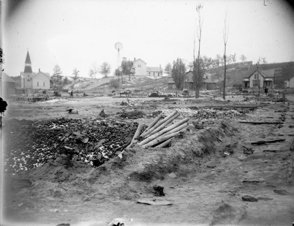 View of pipes in a ditch in front of a church, windmill, and three frame houses.