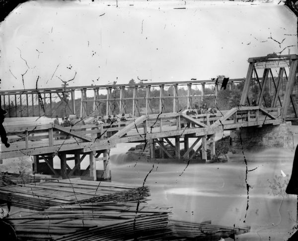View from shoreline of people, men, women and children, gathered in groups on a wooden bridge. A sign in the middle of the bridge reads: "$50 Fine for Cutting, Marking, or Defacing this Bridge. Half the Fine Goes to Complainant." Behind the first bridge is another, higher bridge, perhaps a railroad bridge. In the foreground are stacks of lumber.