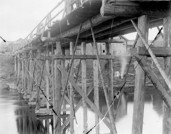 View through the support structure of a trestle bridge. On the shoreline in the background a woman, girl, and another person are standing around what appears to be a campfire. Above them up the steep bank two men stand in front of a large group of log buildings.